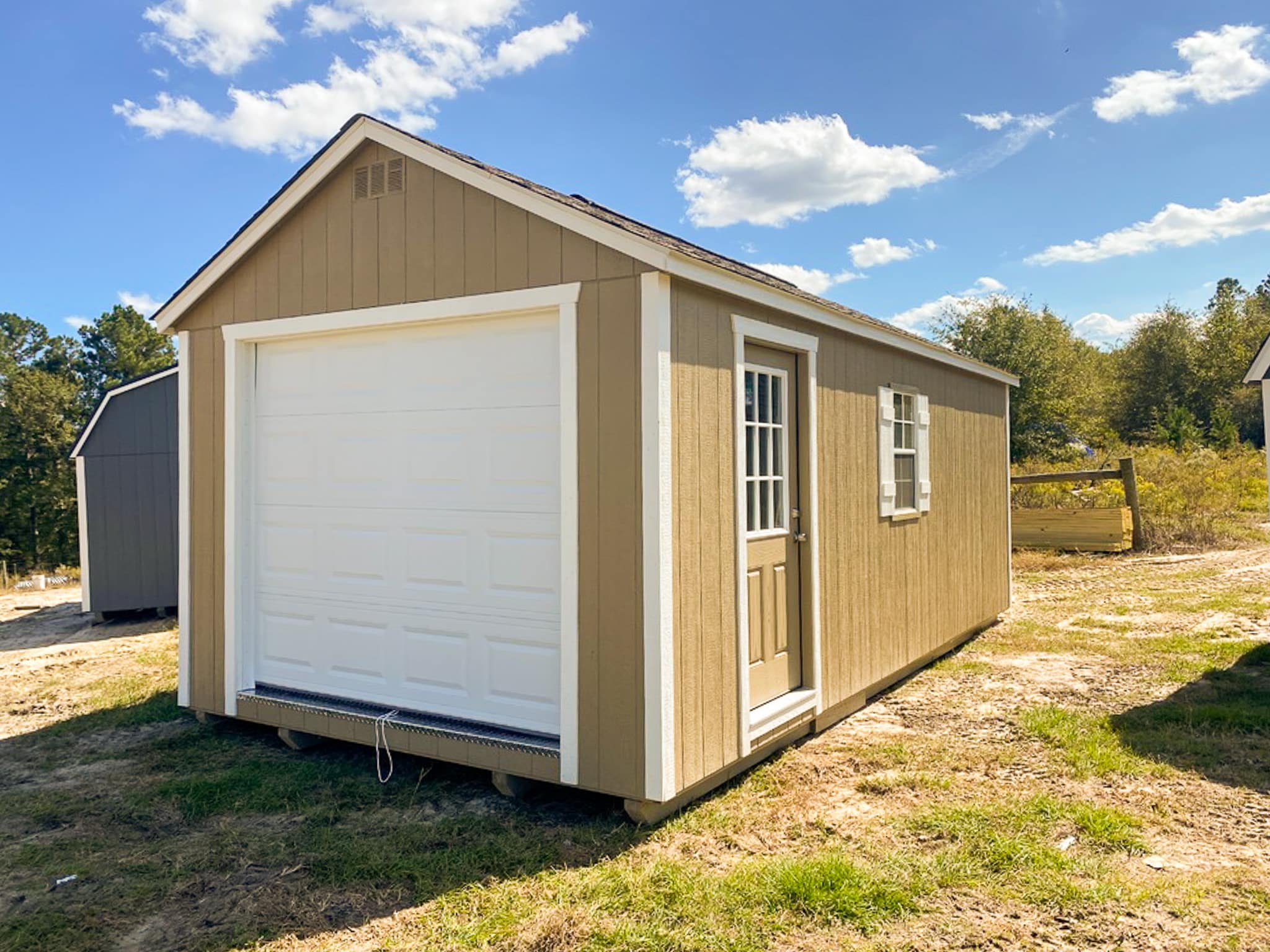 Our 12x24 garage in Jesup GA - Buckskin colored siding with white overhead door with matching trim on corners - Buckskin colored side door inset with window and window with white shutters under weathered wood toned shingled roof - shed sits on patchy grass beneath blue sky dotted with clouds