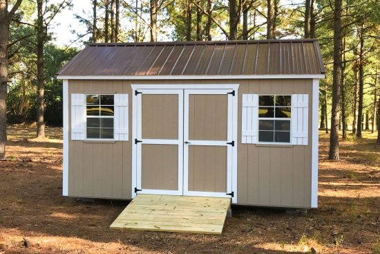 Beige garden shed max with wooden ramp leading to white-trimmed double doors between two windows with white shutters under a brown roof-One of our stylish sheds in Grovetown