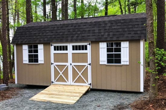 Lofted Barn Max-Tan siding with white-framed windows and double doors under a brown roof-wooden ramp in front-green forest in background-one of our popular sheds in Grovetown