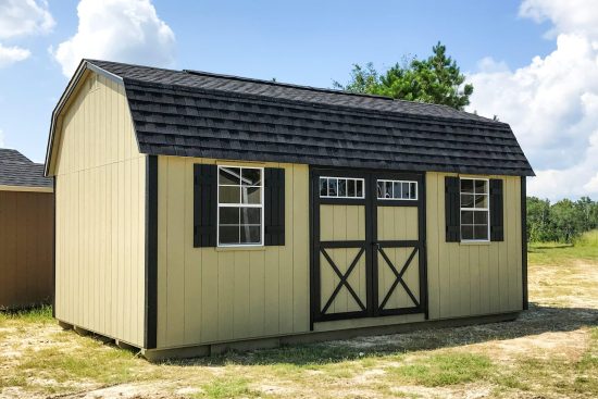 One of our tan lofted barn sheds in Waycross-black trimmed double doors- two windows with black shutters on each side of doors-black barn style roof- some trees in background