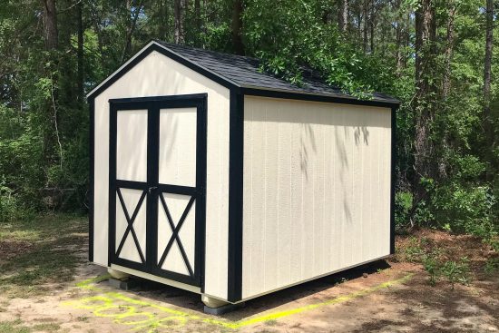 One of our utility sheds in Waycross-white with black trimmed double doors and black roof in front of green wooded area