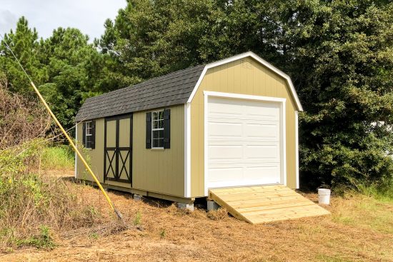 One of our garage sheds in Douglas GA - beige siding with brown-trimmed double doors with matching window shutters and roof white overhead door on with natural wood ramp - lush green trees in background