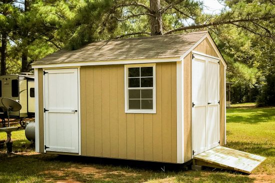 One of our beige utility sheds in Douglas GA - Beige wooden siding with white single and double doors with matching window - Brown shingled roof - green grass and pine tree in background