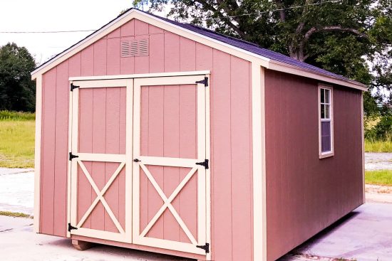 One of our pink utility building sheds in Douglas GA - Pink wooden siding with light pin trimmed double doors and window frame under brown roof - tree in background on the right