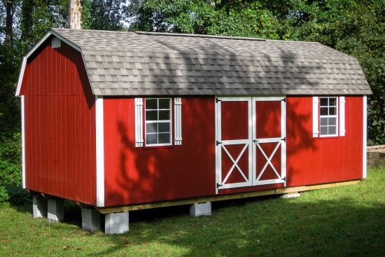 One of our red Lofted Barn Max sheds in Cordele - Red siding with white-trimmed double doors and white windows beneath gray roof - shed on grass in front of green trees