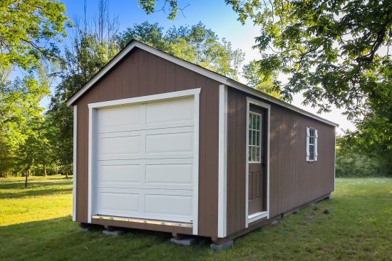 Brown and white garage - Brown painted wood siding with white-trimmed window and windowed door and white overhead door - shed is on grassy field framed by green trees