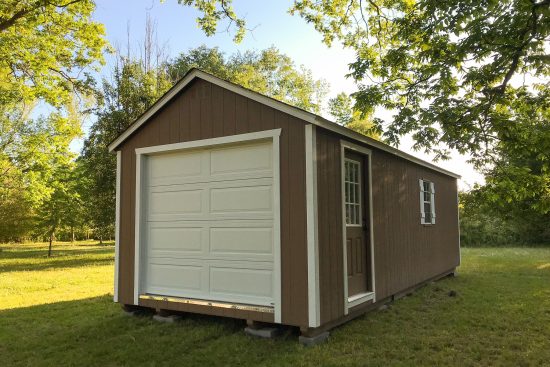 One of our brown garage sheds in Athens GA - Brown painted wood siding with white overhead door and brown side door with window - side window with white shutters - shed on grass tree-lined field