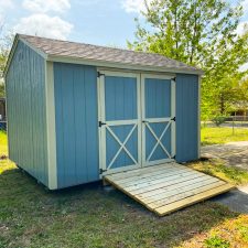 Blue and light silver maple 10x12 Utility Shed in Augusta GA - Blue siding with light silver trimmed double doors with X on lower half - black shingled roof - shed sits on green lawn with green trees in background