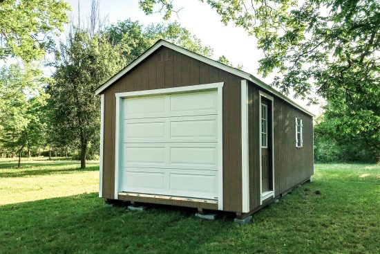 One of our brown and white garage sheds in McDonough GA - Brown wood siding with white overhead door and matching shutters on side window - Single brown entry door with inset window - gable-style roof - garage sits on green grassy area with trees in near distance