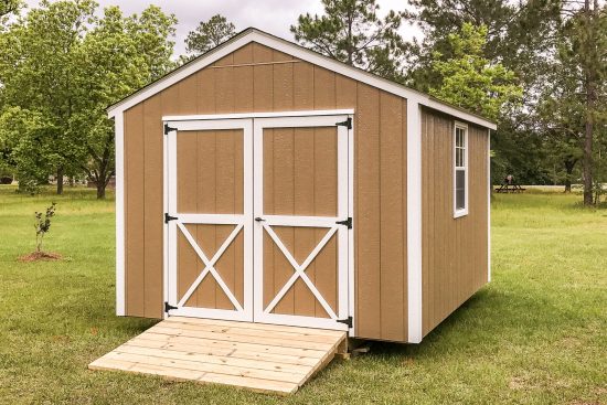 One of our beige Utility Sheds in McDonough GA - beige painted siding with white-trimmed double doors, matching corners and window beneath gable-style roof - shed sits with natural wood ramp on green grass with trees in near distance