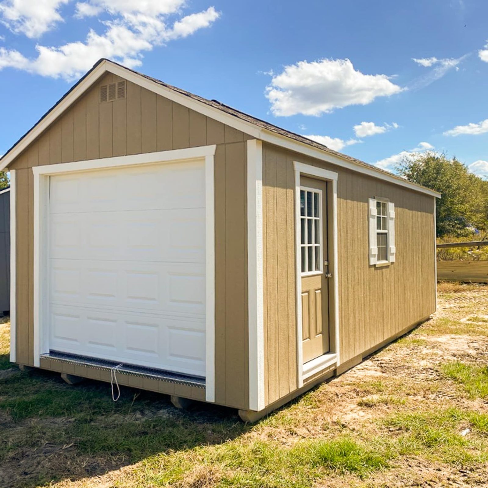 Our 12x24 garage in Jesup GA - Buckskin colored siding with white overhead door with matching trim on corners - Buckskin colored side door inset with window and window with white shutters under weathered wood toned shingled roof - shed sits on patchy grass beneath blue sky dotted with clouds