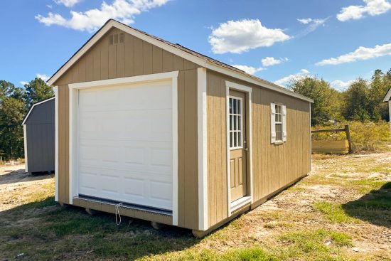 Our 12x24 garage in Jesup GA - Buckskin colored siding with white overhead door with matching trim on corners - Buckskin colored side door inset with window and window with white shutters under weathered wood toned shingled roof - shed sits on patchy grass beneath blue sky dotted with clouds