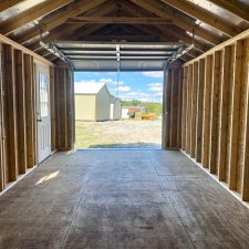 Interior view of 12x24 garage in Jesup GA - unfinished walls exposed beams truss and rafters - window and windowed door on the leftmost wall - open overhead doorway reveals other light-colored buildings nearby beneath blue sky dotted with white clouds