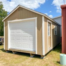 Our 12x24 garage in Jesup GA - Buckskin colored siding with white overhead door with matching trim on corners - Buckskin colored side door inset with window and window with white shutters under weathered wood toned shingled roof - shed sits on patchy grass beneath blue sky dotted with clouds