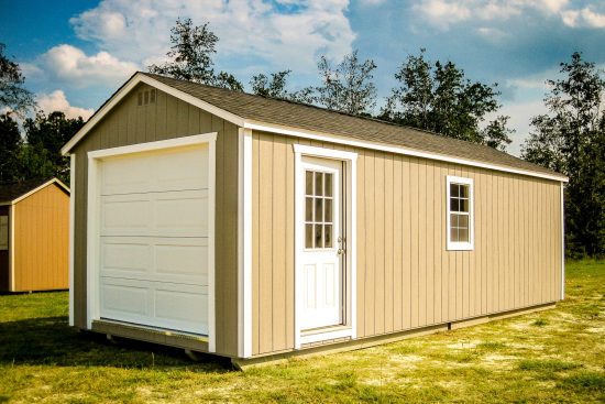 One of our garage sheds in North Augusta SC - Beige painted wood siding with white overhead door on short wall and white-framed window and matching door beneath brown shingled roof - garage sits on patchy yard with treetops visible nearby