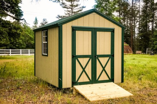 One of our beige utility sheds in North Augusta SC - Beige painted wood siding with dark green trimmed corners and double doors with X detail - green-bordered white-framed window on left side under dark green metal roof - shed sits on grassy yard with white- fenced tree-lined border