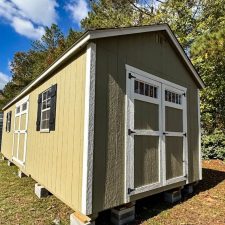 Corner perspective of a beige 12x28 Garden Shed Max with white double doors and black shutters in front of trees beneath blue partly cloudy sky