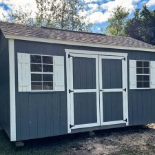 Gray 10x16 Garden Shed Max with white-trimmed double doors and shutters in fenced back yard beneath blue partly cloudy sky