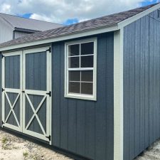 Side view of a gray 12x16 Utility shed with silver maple trim and barn-style doors ( with X detail )on a gravel foundation - stands on lot beneath cloudy blue sky