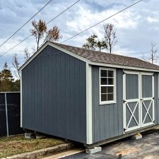 Side view of a gray 12x16 Utility shed with silver maple trim and barn-style doors ( with X detail )on a gravel foundation on a cloudy day