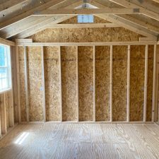 Interior view of a wooden 12x16 Utility Shed with exposed studs, beams and rafters. Natural light filtering in from window.