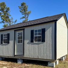 Beige 12x20 Garden Shed Max with black shutters and a single-entry door elevated on sturdy blockson large yard beneath blue sky