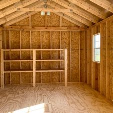 Interior view of 12x20 Garden Shed Max with light filtering through window and 9 lite door, highlighting exposed wooden beams, studs and rafters