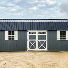 A gray 12x28 Lofted Barn Max with a black metal roof, white trim, double barn-style doors, and windows with white shutters, set against a cloudy sky.