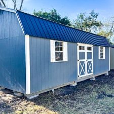 A gray 12x28 lofted barn max with a black metal roof, white trim, and double barn-style doors, elevated on concrete blocks, set in a wooded outdoor area.