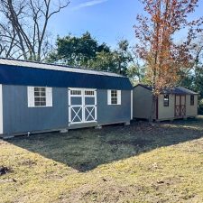 Gray 12x28 Lofted Barn Max beside beige and red 12x24 utility shed in yard among trees