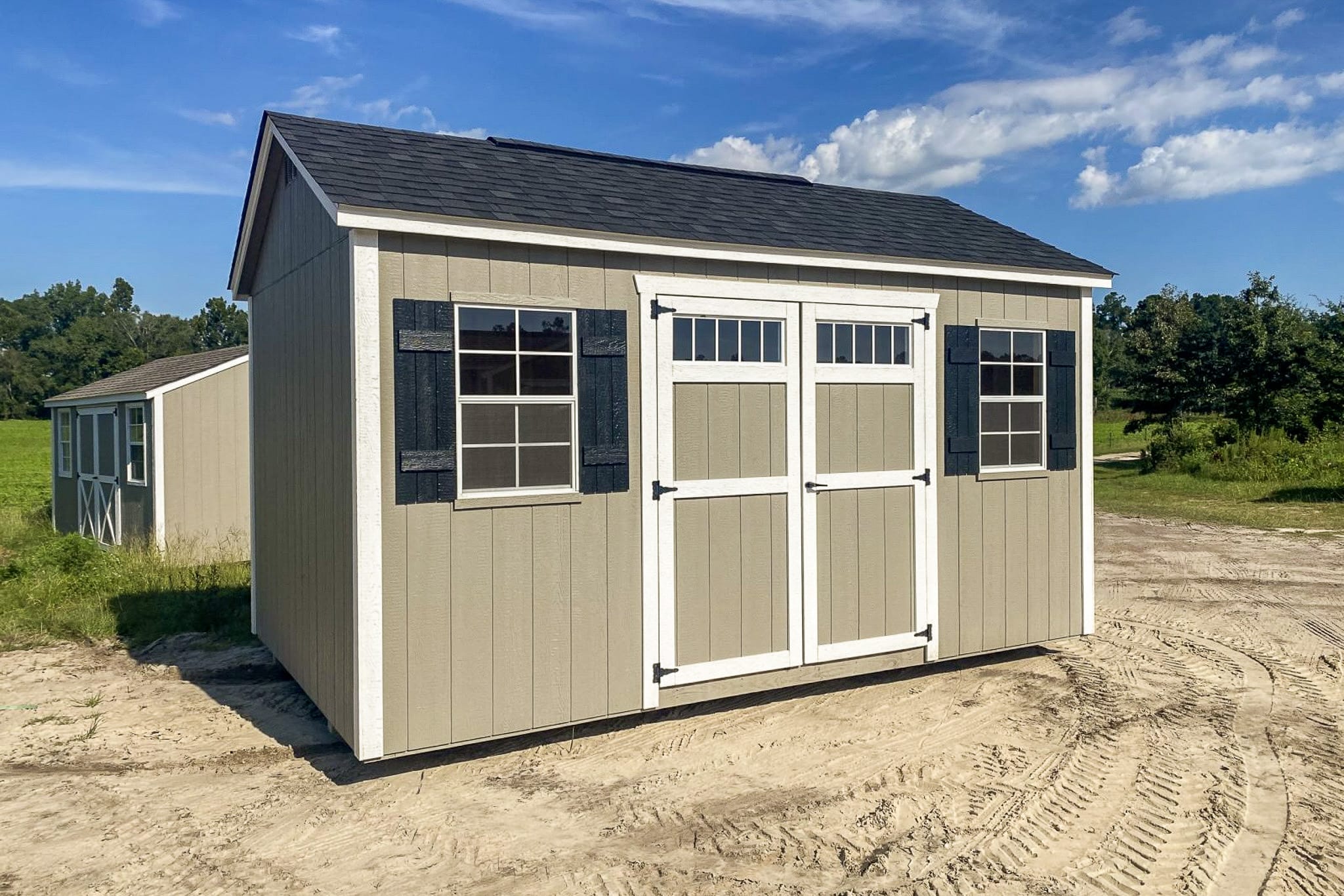 One of our Garden Shed Max Sheds in McDonough GA - Tan painted wood siding with white trimmed corners and double doors between two white-framed windows with black shutters under black shingled roof - shed sits on sandy area with visible green grass and trees in the near distance beneath blue sky with a few white clouds