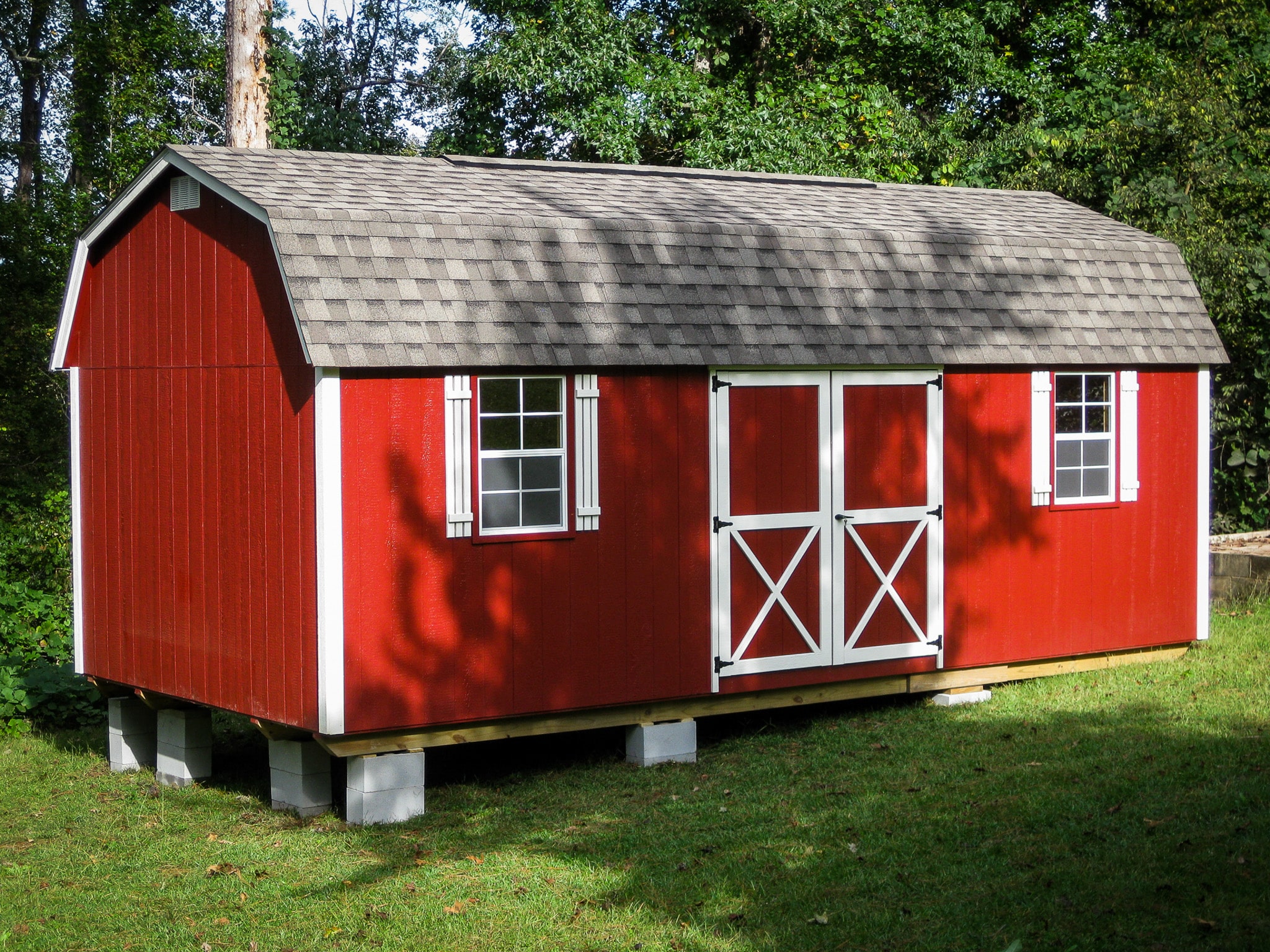 One of our red Lofted Barn Max sheds in Cordele - Red siding with white-trimmed double doors and white windows beneath gray roof - shed on grass in front of green trees