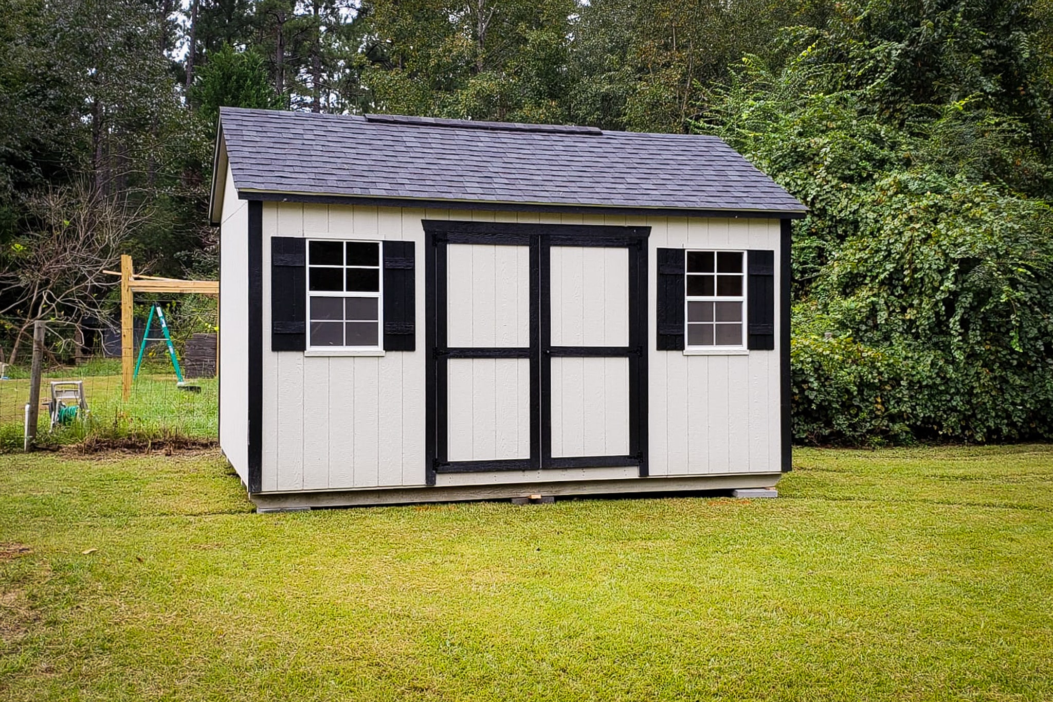 Black and white Garden Shed Max - White wood siding with black trimmed double doors windows and corners under a matching black shingled roof - shed on green grass in front of lush green trees